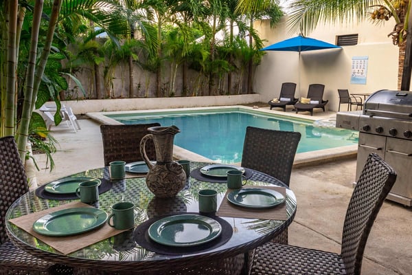 Courtyard with dining table set for meal, swimming pool, blue umbrella, lounge chairs, and gas grill surrounded by palm trees