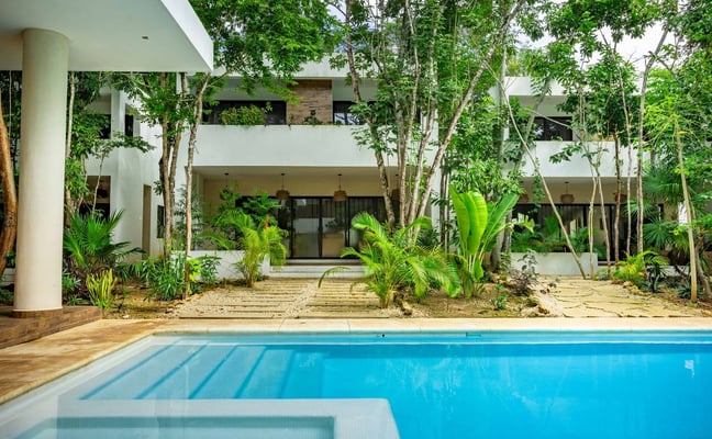 Tropical courtyard with turquoise pool, thatched-roof stone buildings, palm trees, and lounge chair