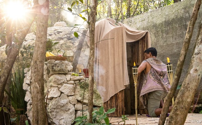 Shaman kneeling at the entrance of a stone temazcal dome in the jungle at sunrise