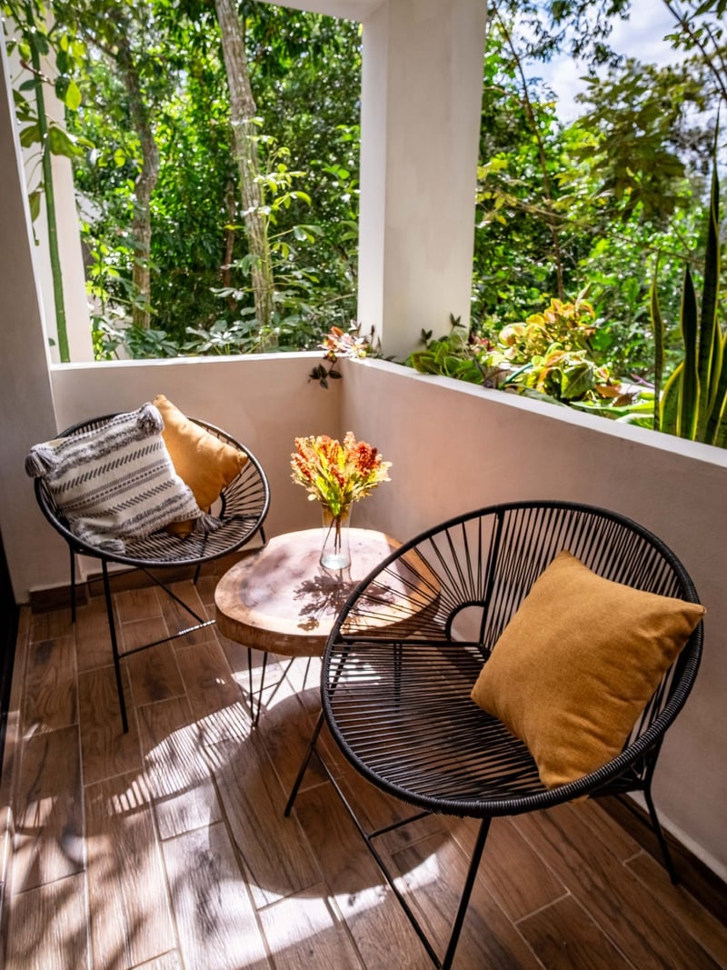 Balcony with black wicker chairs, small table, and orange cushions, overlooking lush green foliage.