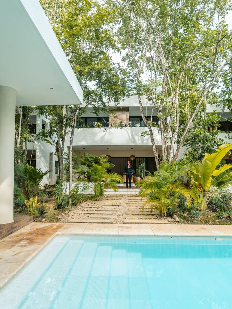 Pool with turquoise water leads to a modern white building. Green plants and trees surround.