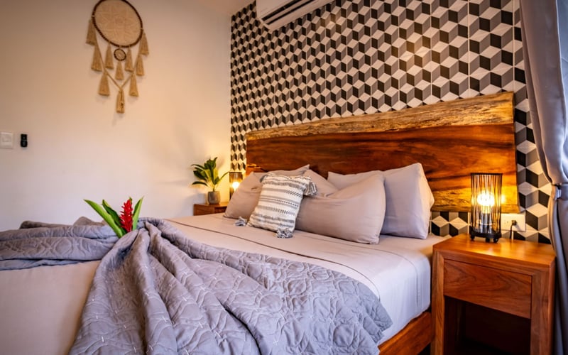 Bedroom with wooden bed frame, patterned wallpaper, dream catcher, and bedside table with lamp.