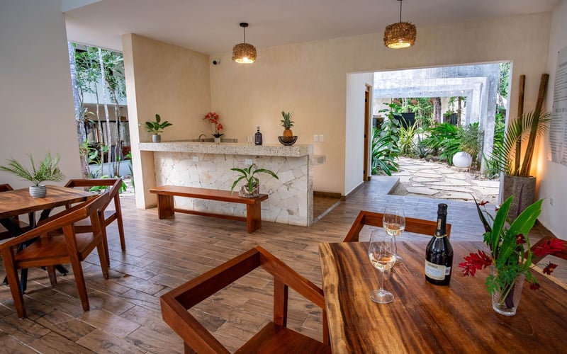 Restaurant interior with wooden tables and chairs, a bar, and an open doorway to a garden.